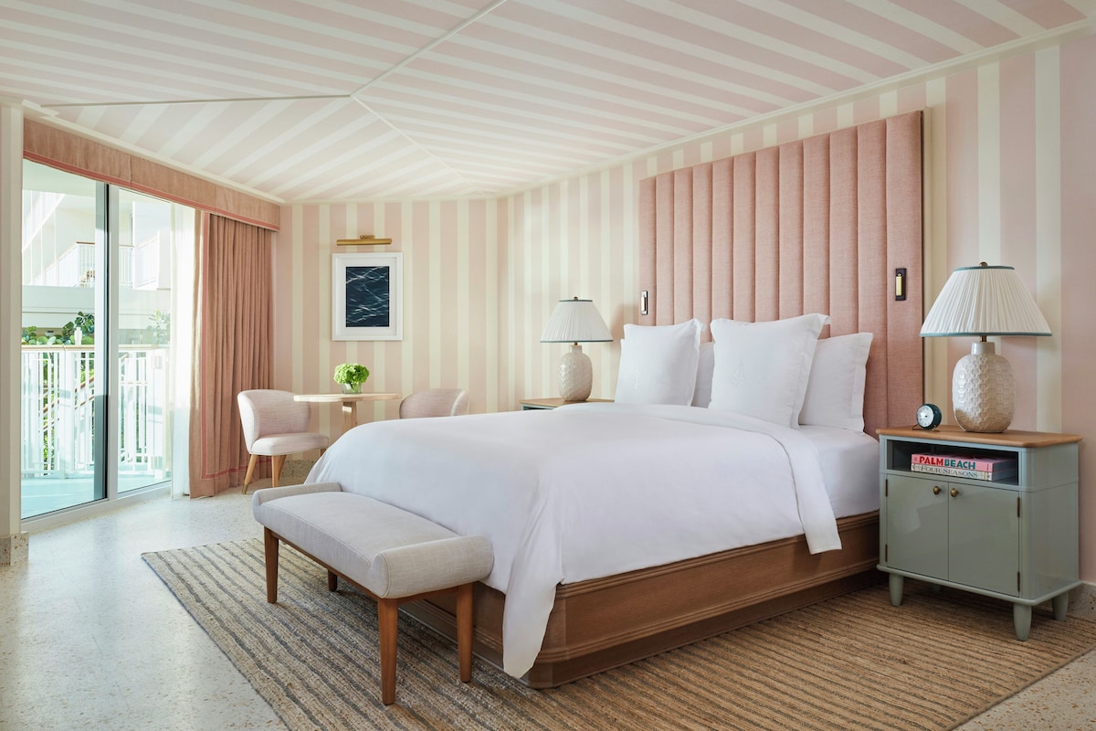 A hotel room with a large bed, bedside lamps, a small table with a plant, a bench, and a view of the outdoors.