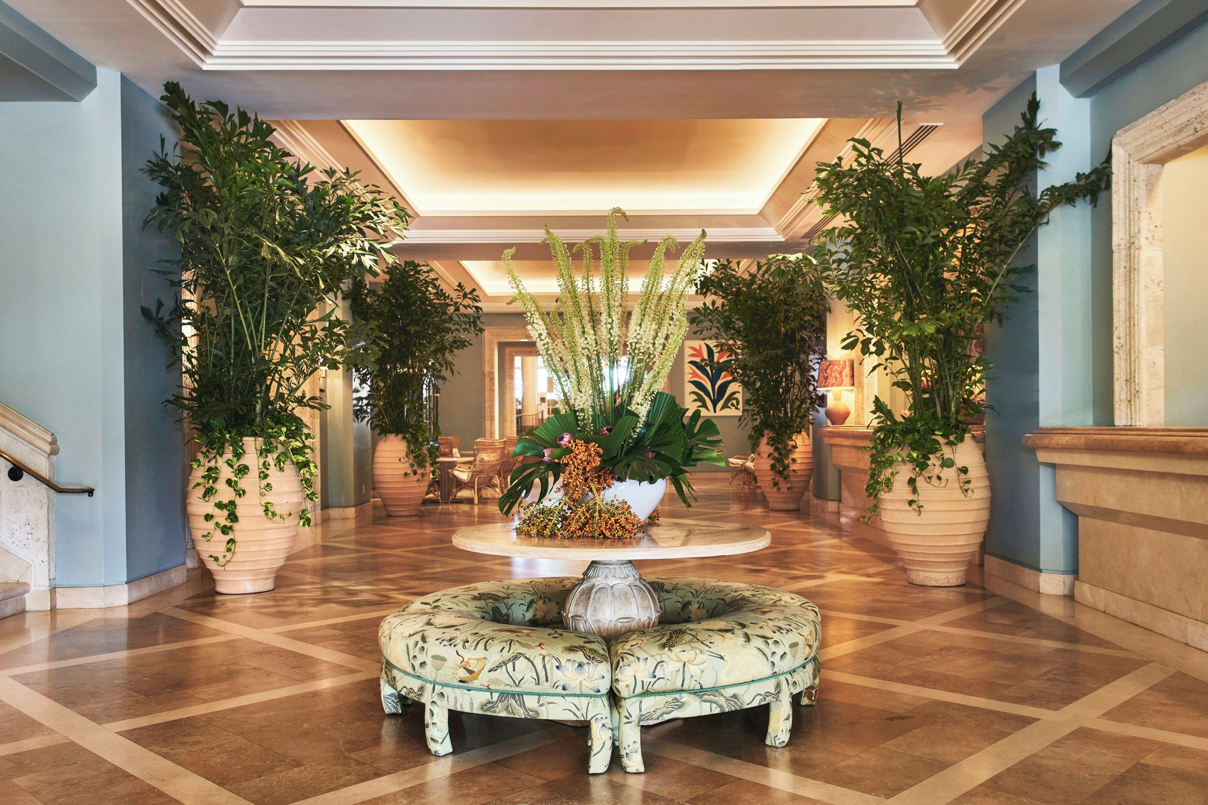 A lobby with a central round table surrounded by potted plants, a patterned bench, and a classical architectural design.