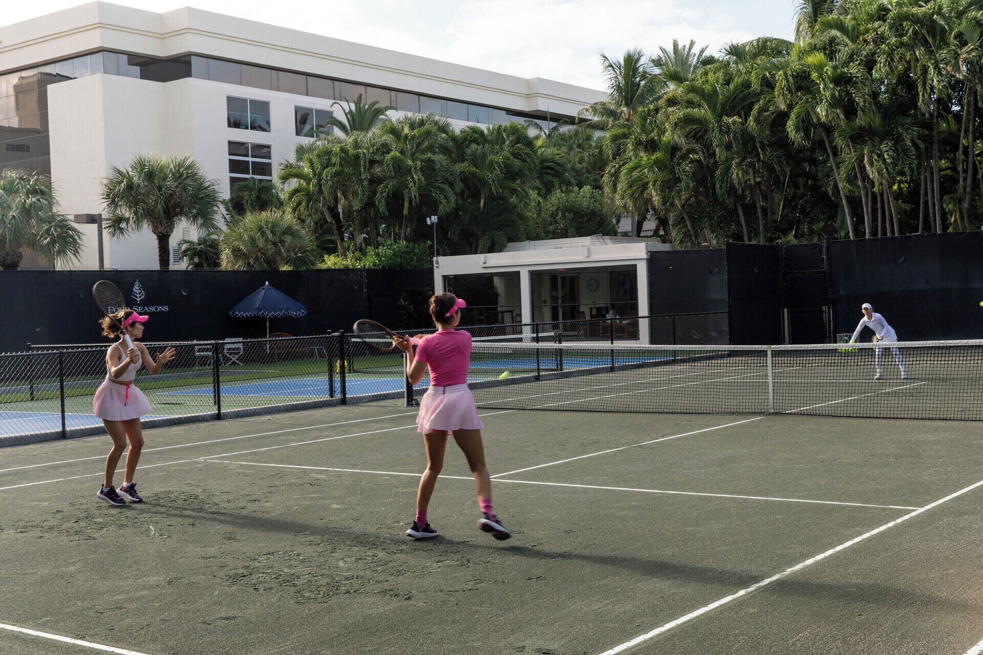 A tennis court with two players in pink outfits, surrounded by palm trees and a building in the background.