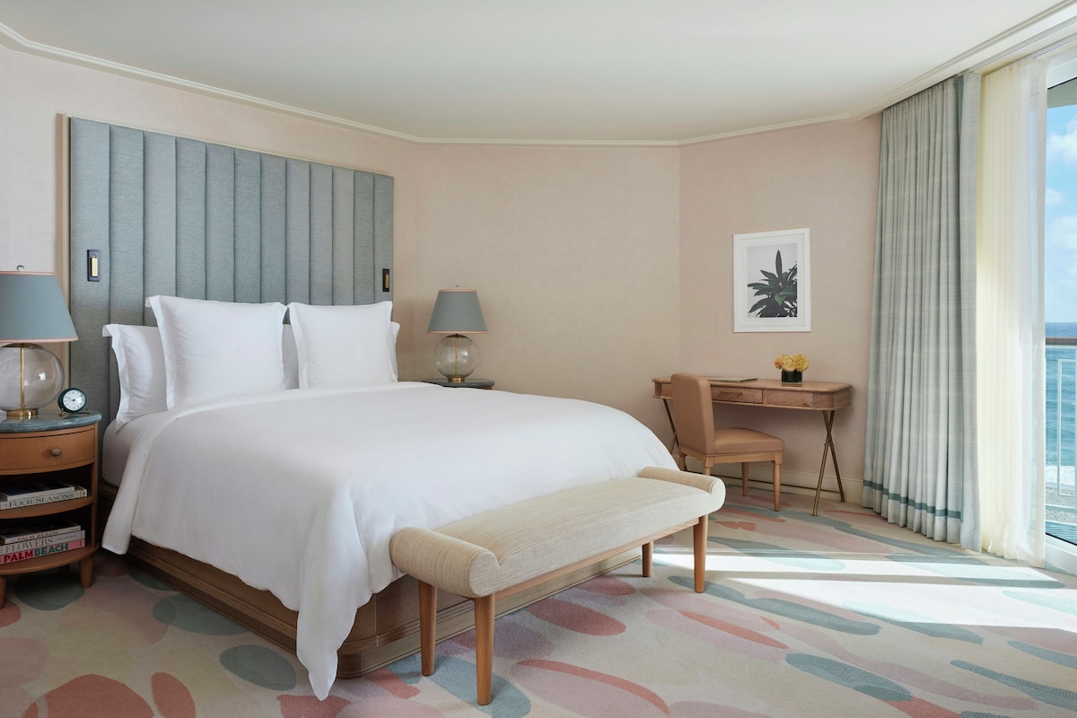 A hotel room with a large bed, two bedside tables with lamps, a bench, a desk with a chair, and a view of the sea.