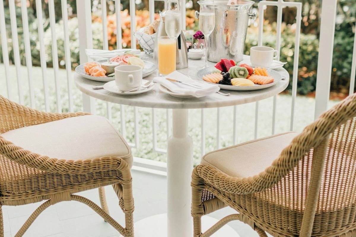 A round table with a white pedestal base, surrounded by wicker chairs with cushions. The table is set with plates of food, glasses, and a bottle of orange juice.