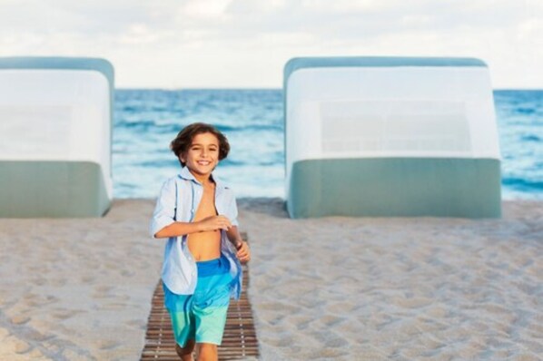 A beach with two white lounge chairs and a person walking between them.
