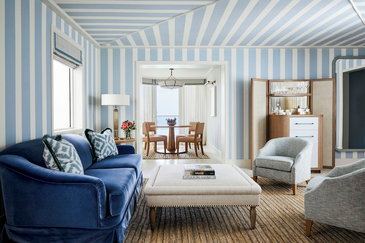 A room with striped wallpaper, a blue sofa, a grey armchair, a wooden coffee table, and a dining area with a chandelier.
