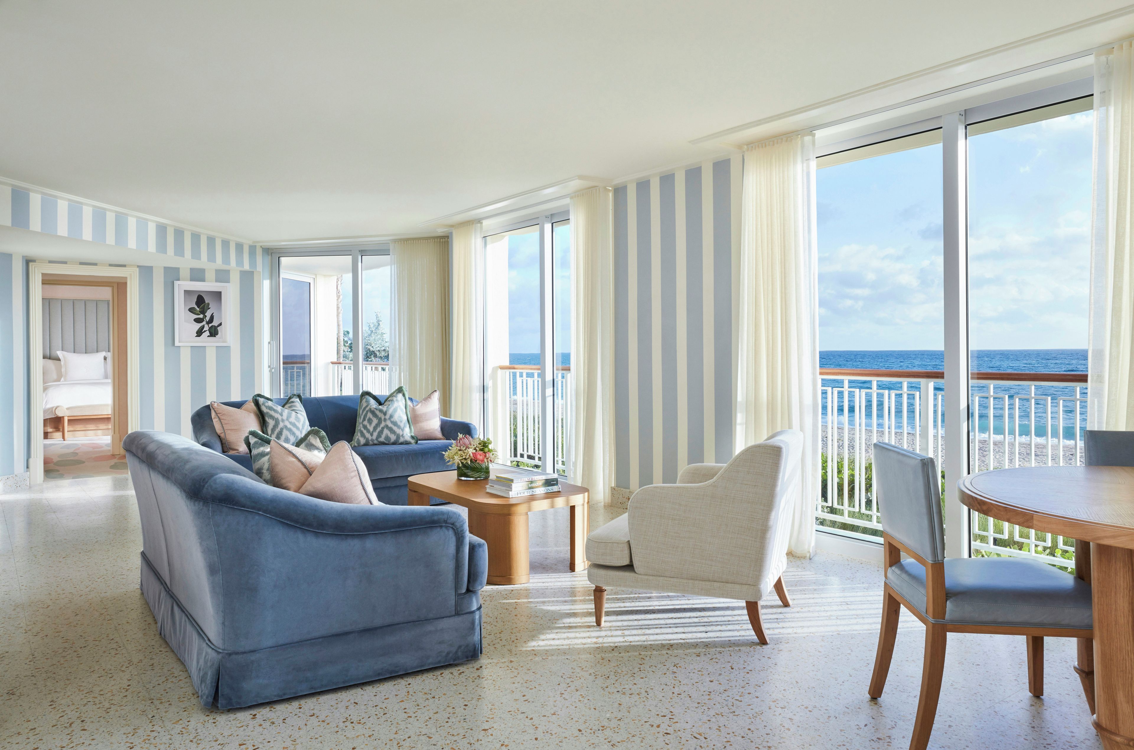 A spacious living room with a blue sofa, a white armchair, and a wooden coffee table. A large window offers a view of the ocean.