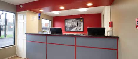 Una zona de recepción con mostrador rojo y gris, dos computadoras, un detector de incendios y una ventana con vistas a la calle.