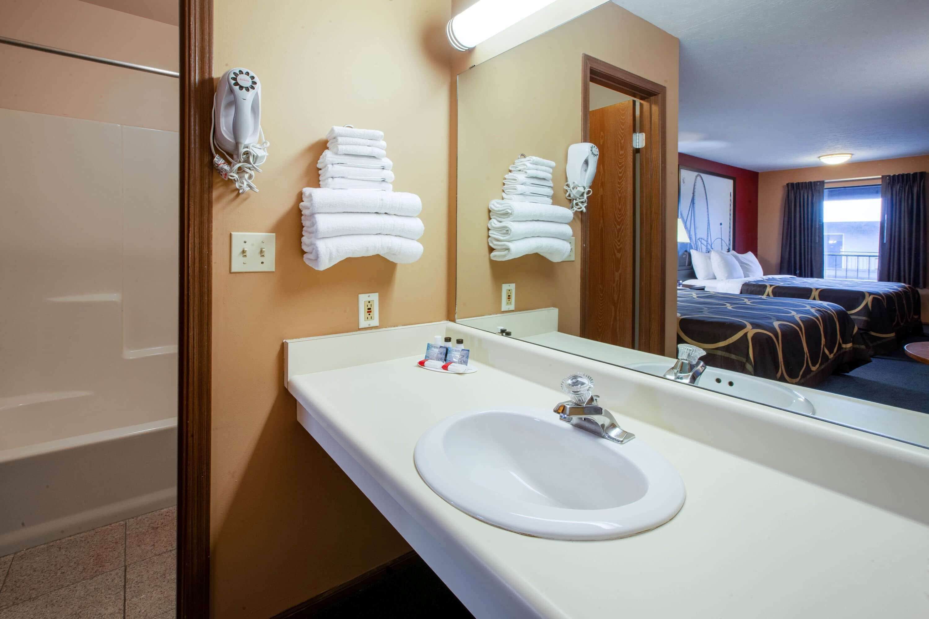 A hotel bathroom with a sink, mirror, towel rack, and a bed visible in the reflection.