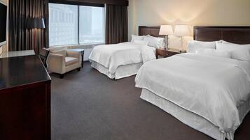 Quarto de hotel com duas camas, uma escrivaninha, uma cadeira e uma janela grande com cortinas.