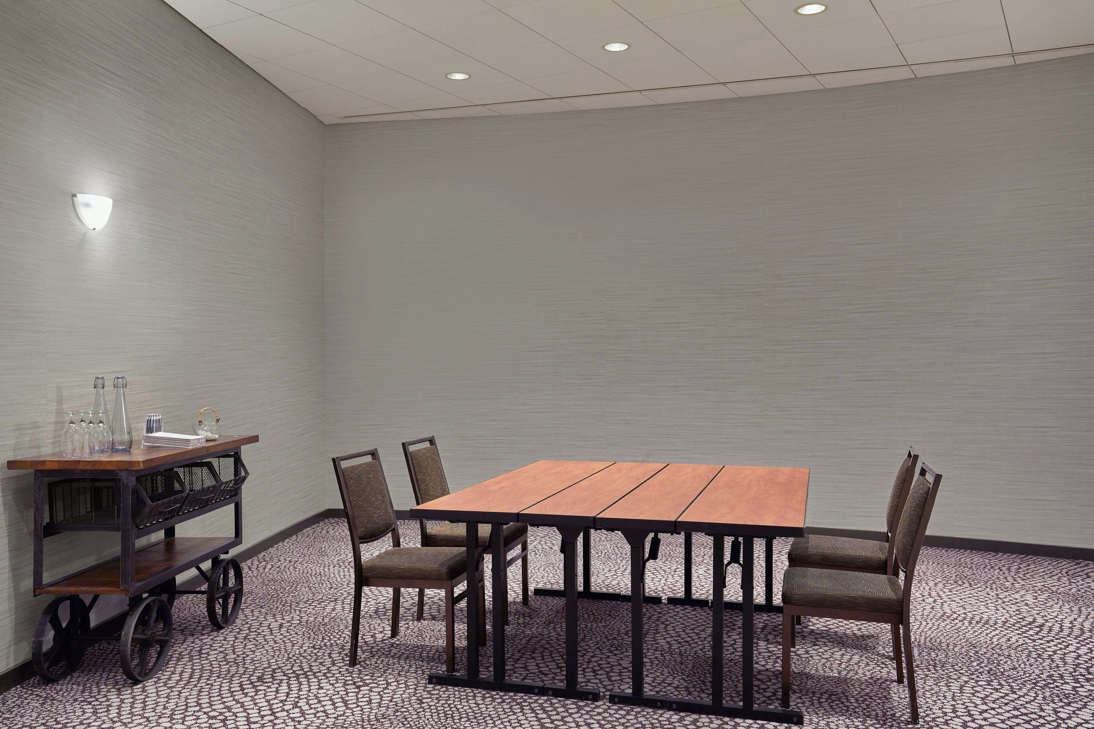 A conference room with a wooden table and four chairs, a small cart with glassware, and a wall-mounted light fixture.