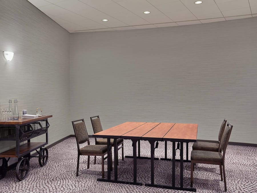 A conference room with a wooden table and four chairs, a small cart with glassware, and a wall-mounted light fixture.
