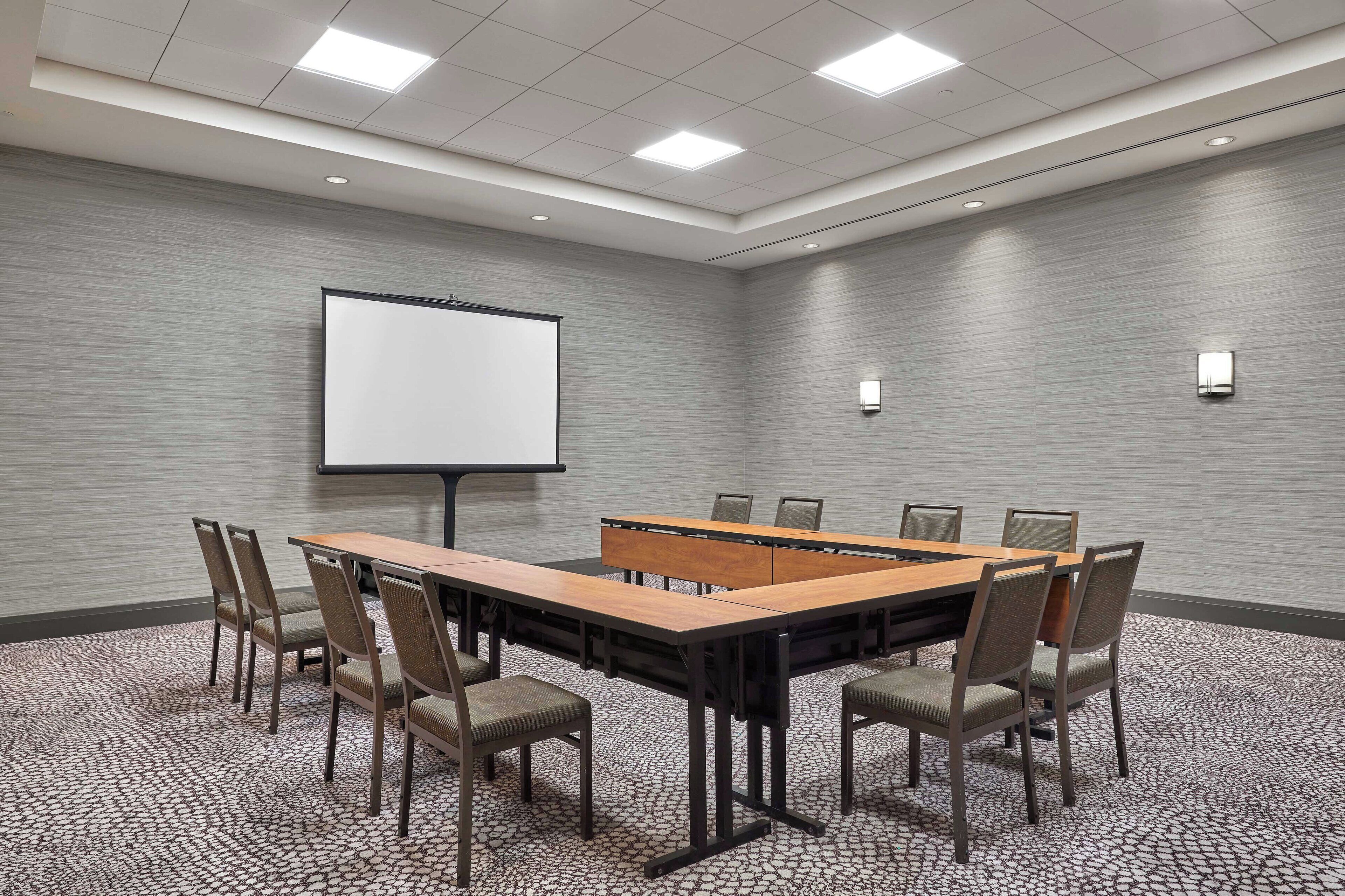 Une salle de conférence avec une longue table, six chaises, un écran de projection et un sol recouvert de moquette.