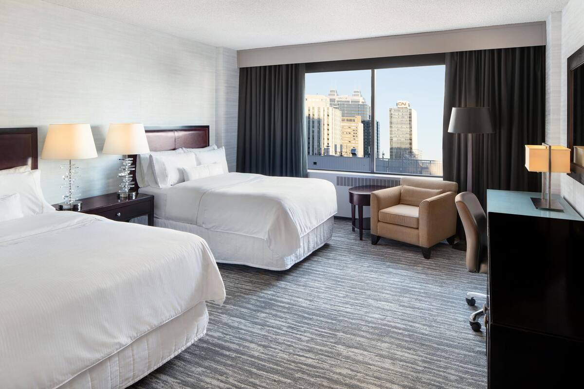 A hotel room with two beds, a desk, a chair, and a large window with a city view.