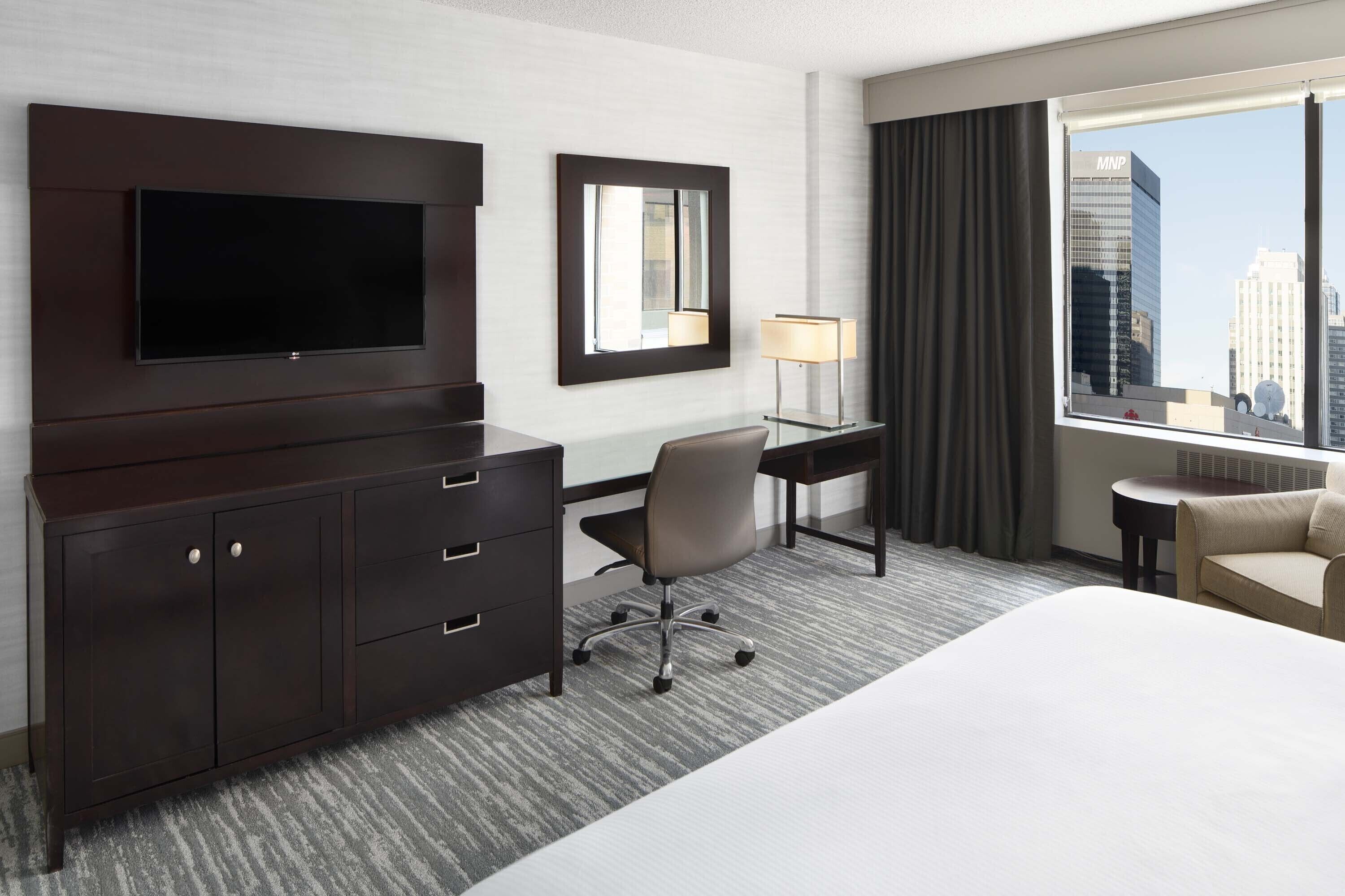 A hotel room with a bed, a desk, a chair, a TV, a dresser, a lamp, and a window with a city view.