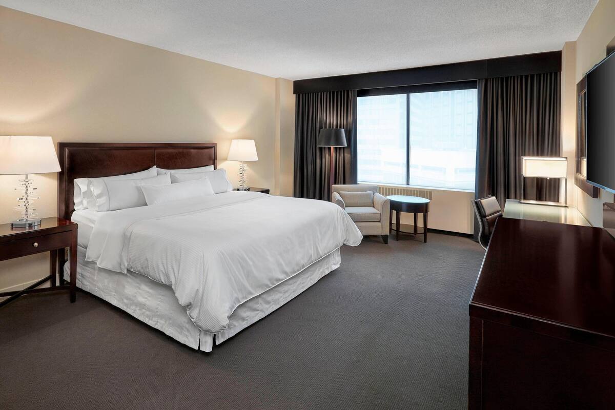A hotel room with a large bed, two bedside tables with lamps, a desk, a chair, and a TV.