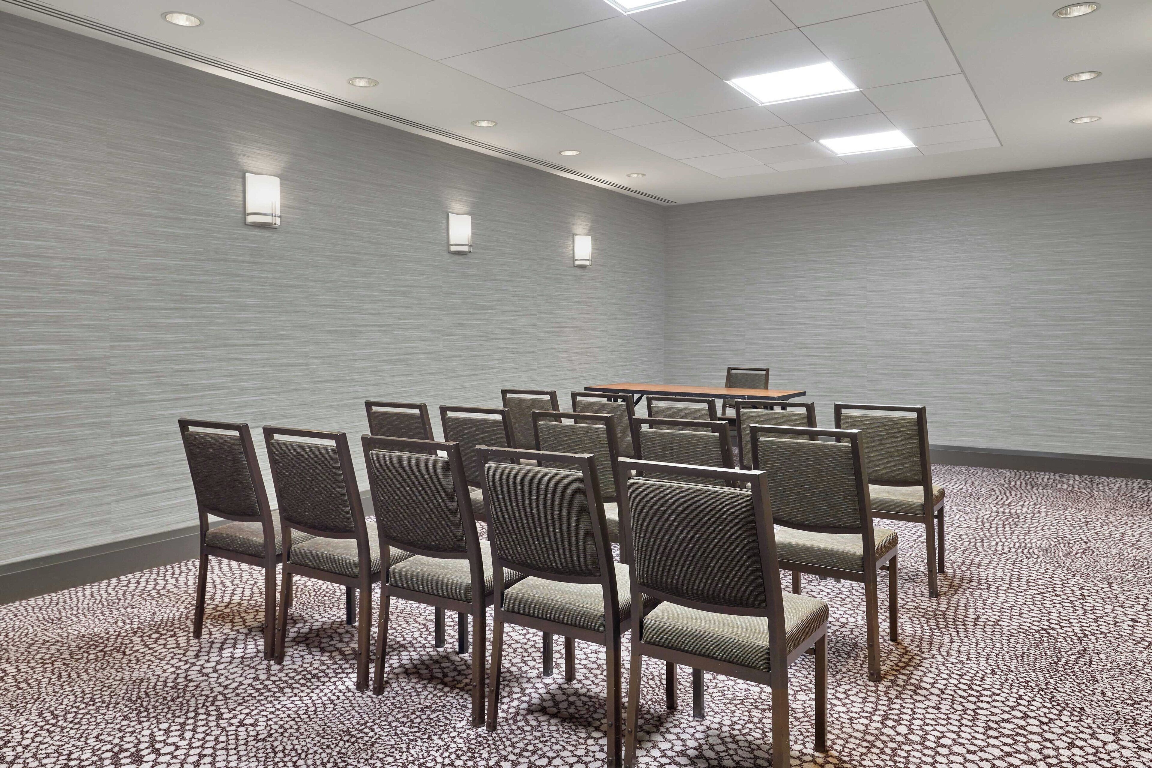 Un salón de conferencias con una mesa rectangular rodeada de sillas, una alfombra estampada y una pared con textura.