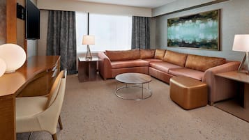 Una habitación de hotel moderna con un sofá modular, una mesa de centro redonda, un escritorio con silla, un televisor de pantalla plana montado en la pared y un cuadro abstracto grande.
