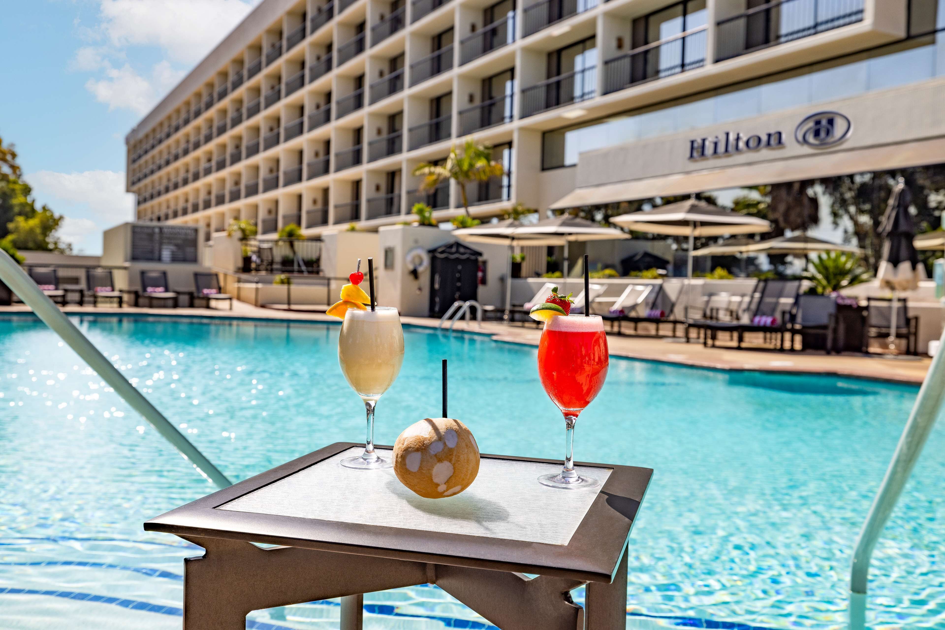 Ein Tisch am Pool mit Getränken und einer Kokosnuss, im Hintergrund ein Hilton-Hotelgebäude sowie Liegestühle und Sonnenschirme.