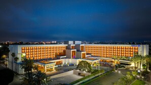 Un hotel moderno con una fachada amplia e iluminada, un estacionamiento espacioso y vistas del horizonte urbano nocturno.