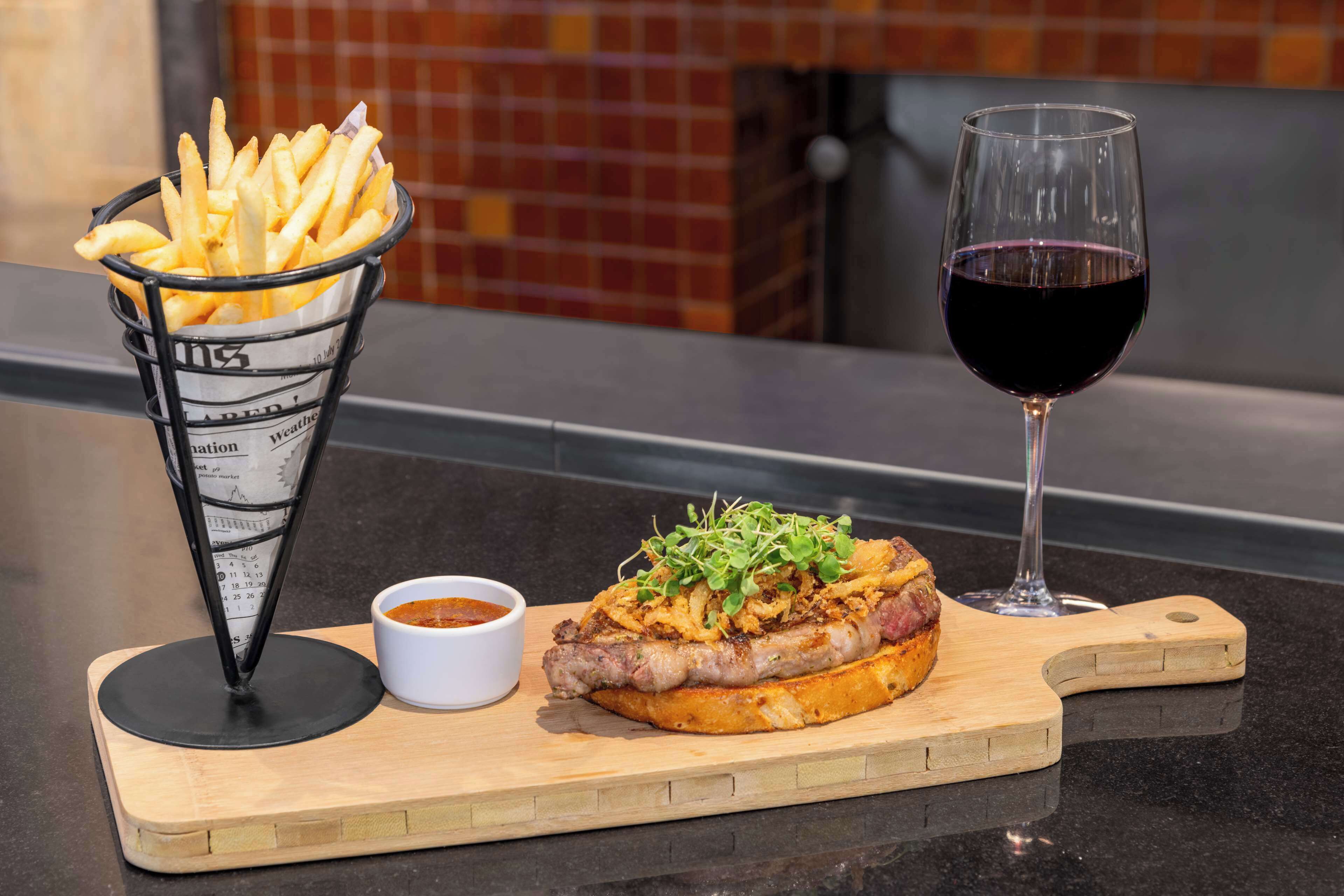 Un repas composé d’un sandwich à la viande et aux légumes, d’un panier de frites, d’un verre de vin rouge et d’un petit bol de sauce, le tout présenté sur une planche en bois.