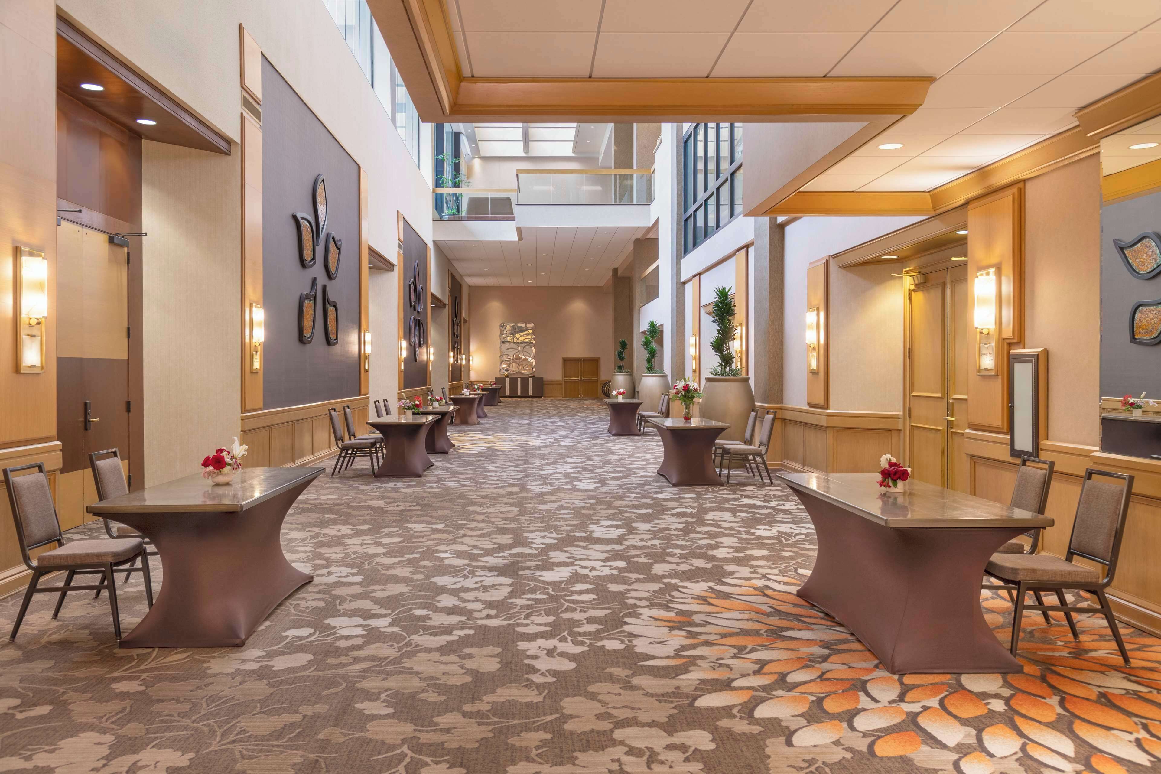 A hotel lobby with a central table and chairs, a patterned carpet, and a high ceiling with skylights.