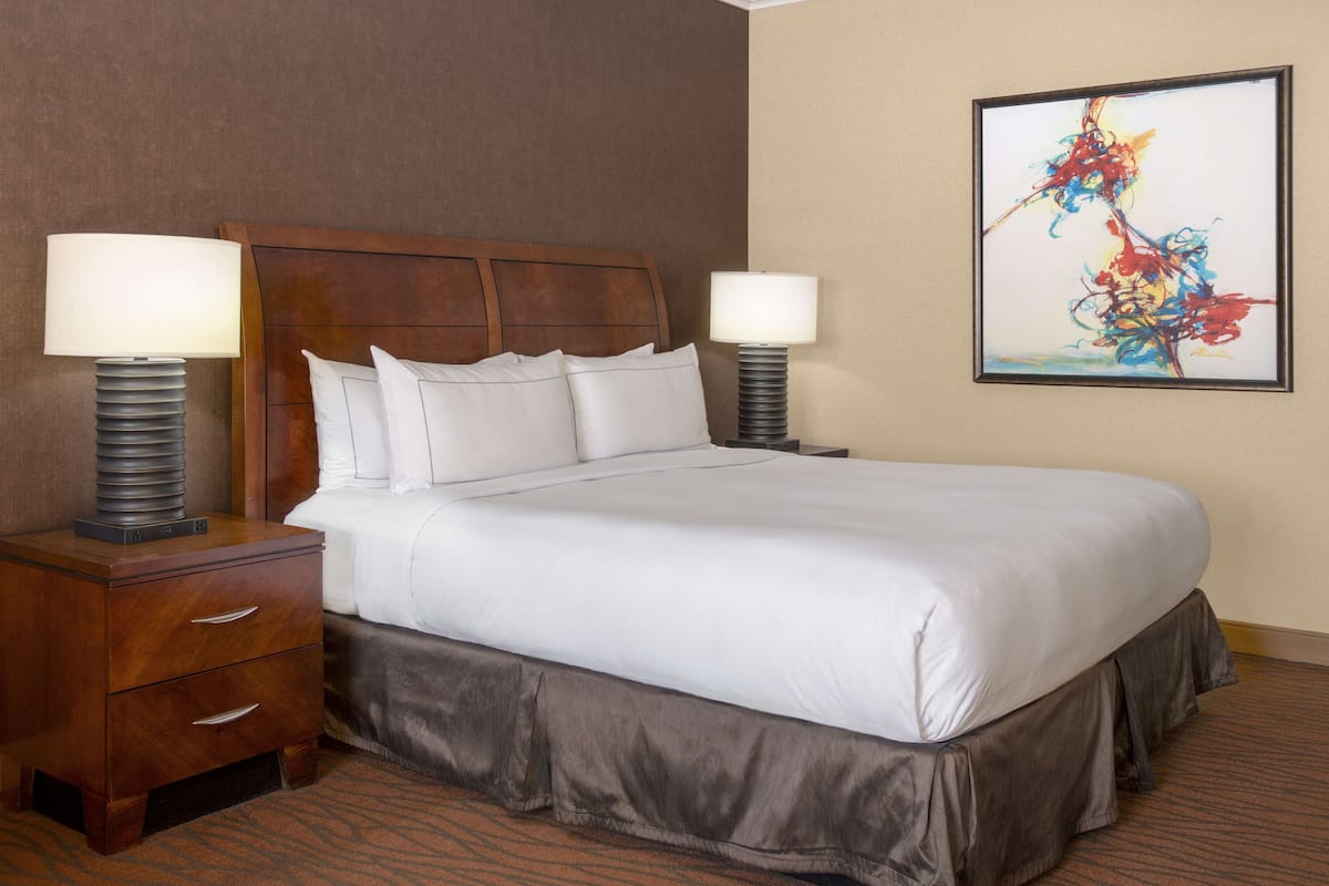 Quarto de hotel com uma cama grande, duas mesas de cabeceira, um painel de cabeceira e um quadro abstrato emoldurado na parede.