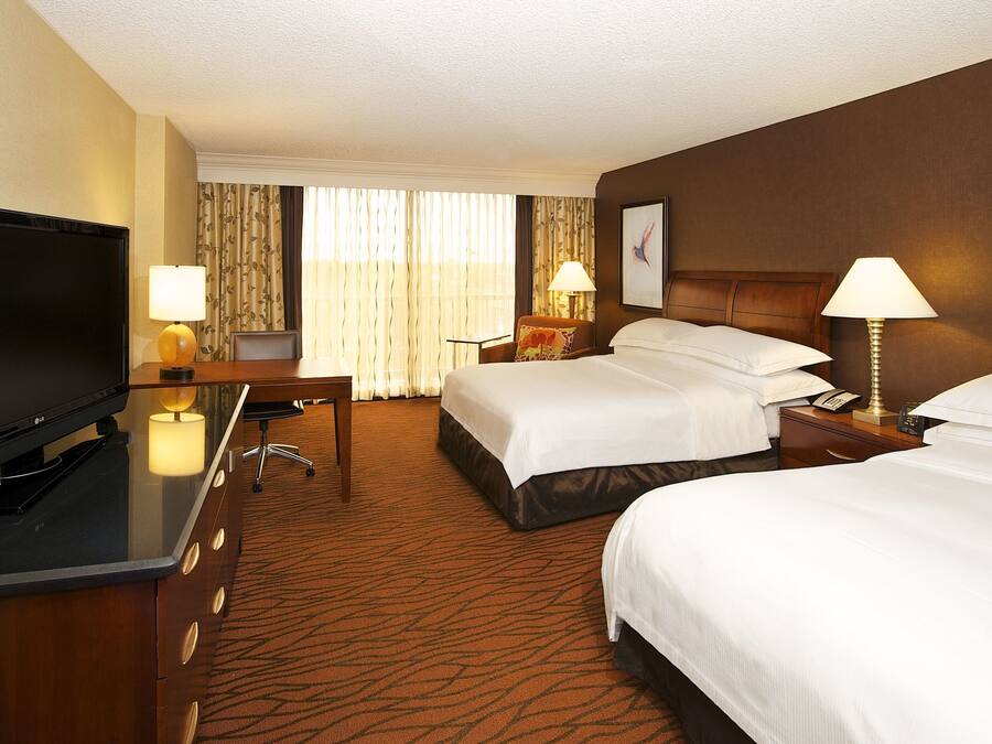 A hotel room with a flat-screen TV, two beds, a desk with a chair, lamps, and a large window with curtains.