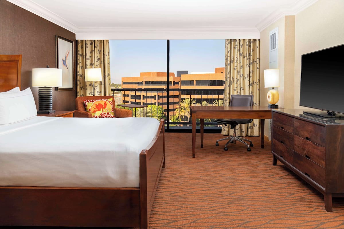 Quarto de hotel com cama, escrivaninha, televisão e vista para edifícios através de uma grande janela.