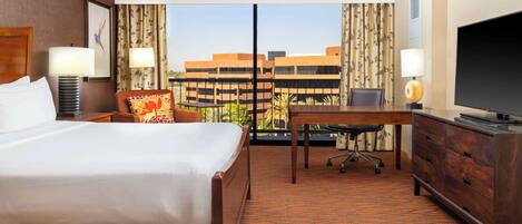 Habitación de hotel con cama, escritorio, televisor y vistas a edificios a través de un ventanal grande.