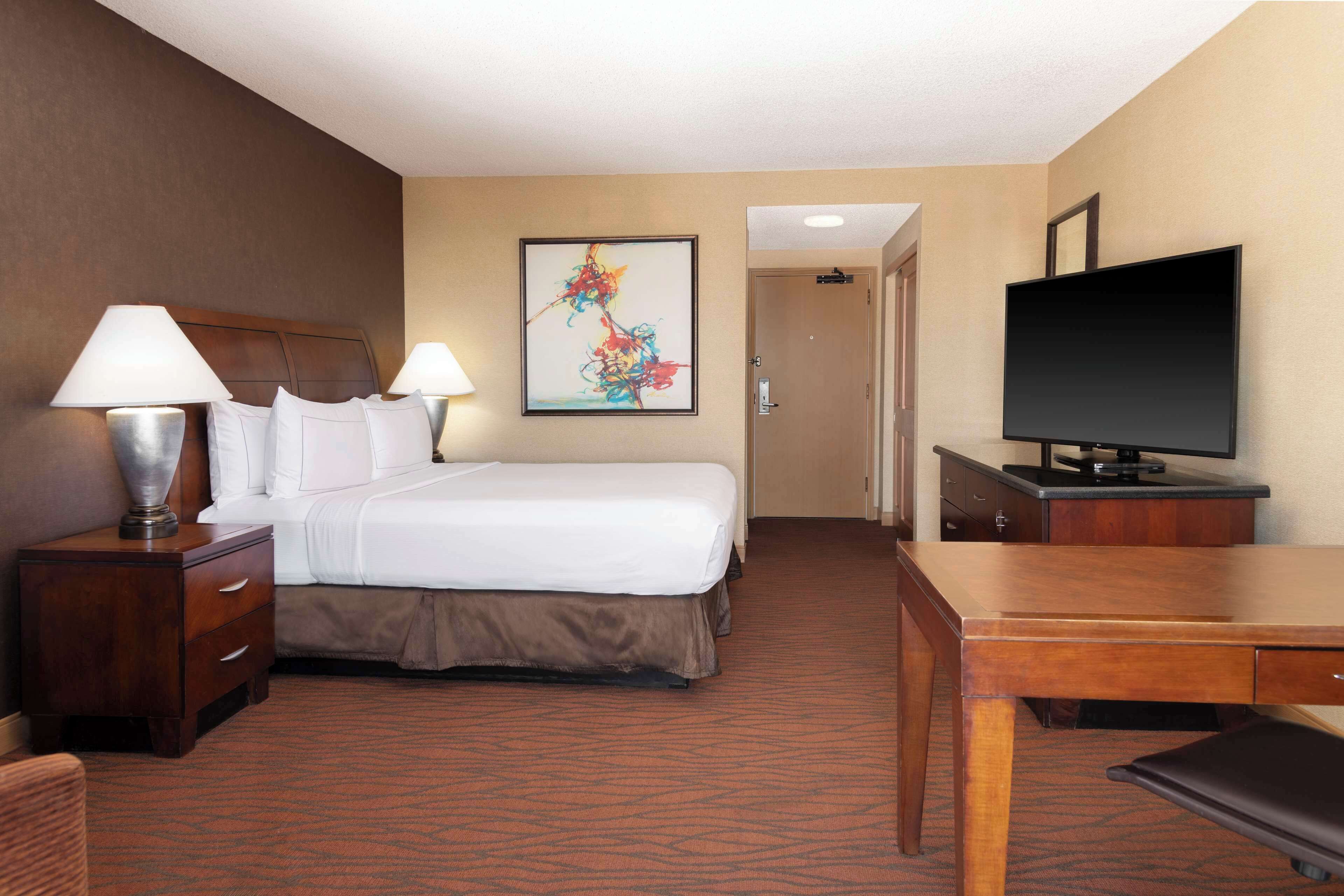 A hotel room with a bed, two bedside lamps, a television, a desk, and a door leading to a hallway.