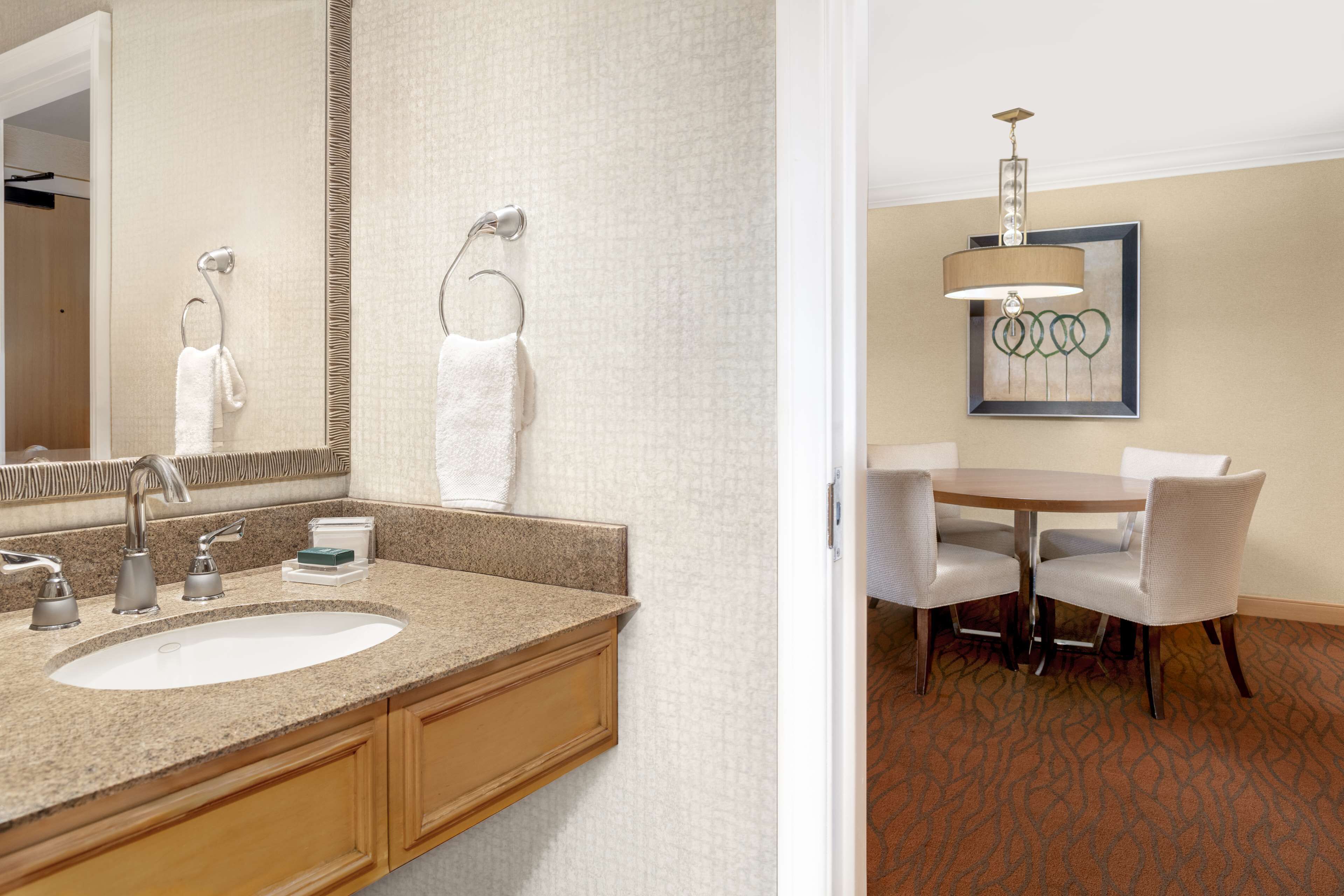 A hotel bathroom with a sink, mirror, towel hooks, and a dining area with a table and chairs.