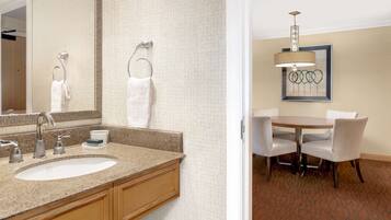 A hotel bathroom with a sink, mirror, towel hooks, and a dining area with a table and chairs.