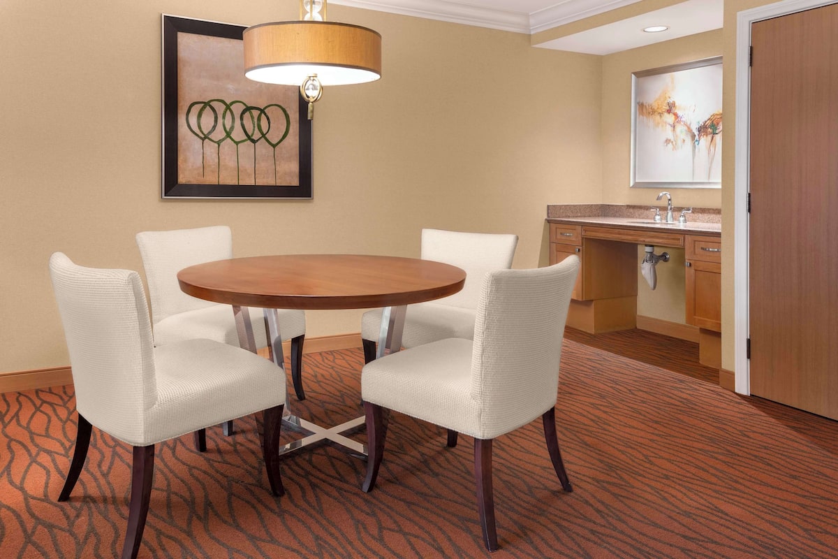 Quarto de hotel com uma mesa de jantar redonda e quatro cadeiras brancas, um armário de madeira com pia e duas obras de arte emolduradas nas paredes.