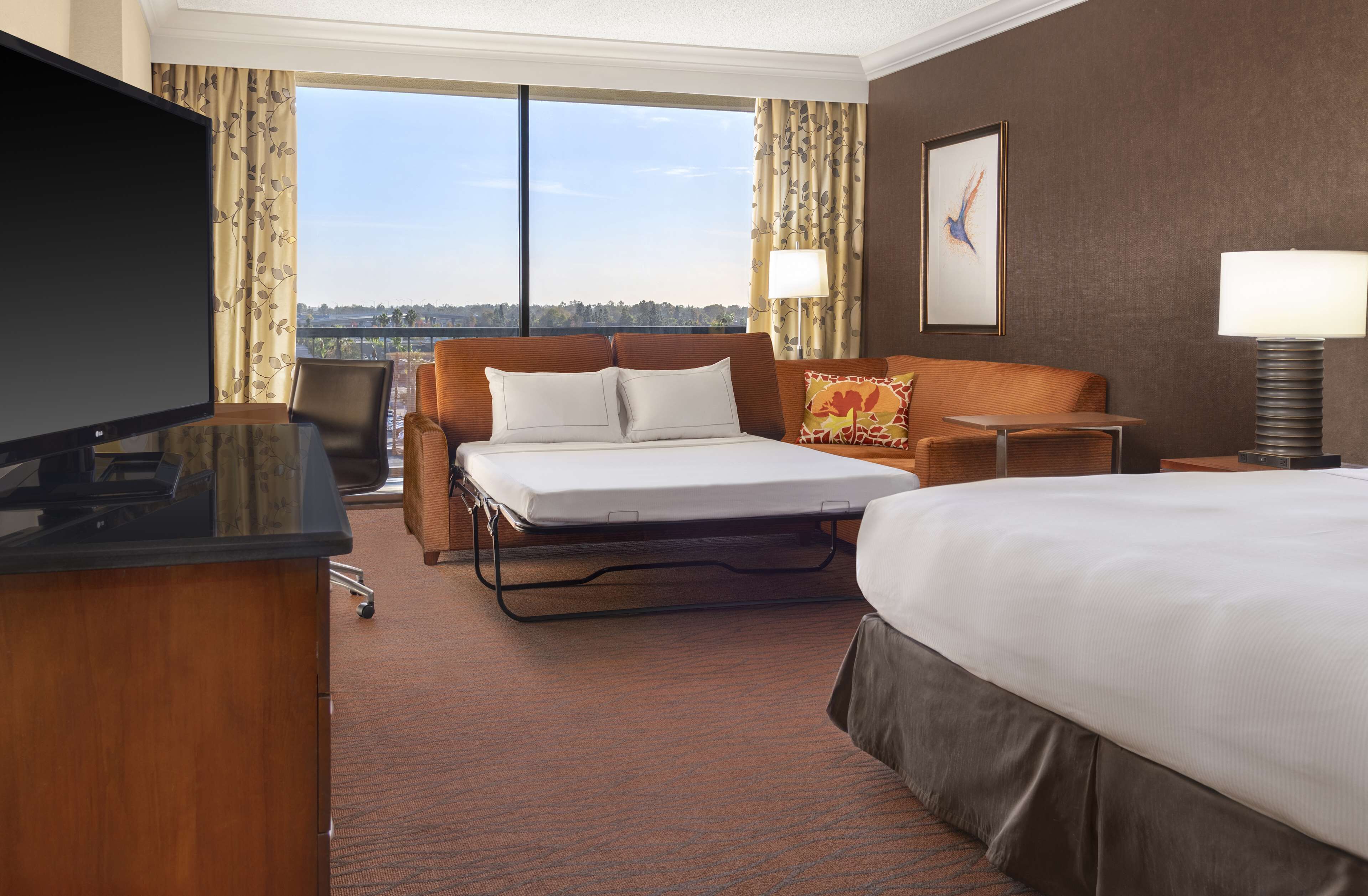 A hotel room with a bed, a desk with a TV, a chair, a small table, and a view of the outdoors.