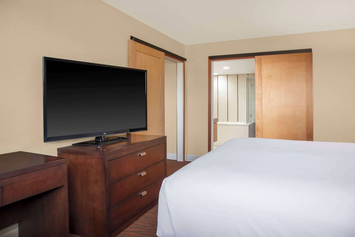 Quarto de hotel com cama, televisão em cómoda de madeira, espelho e porta deslizante para uma casa de banho.