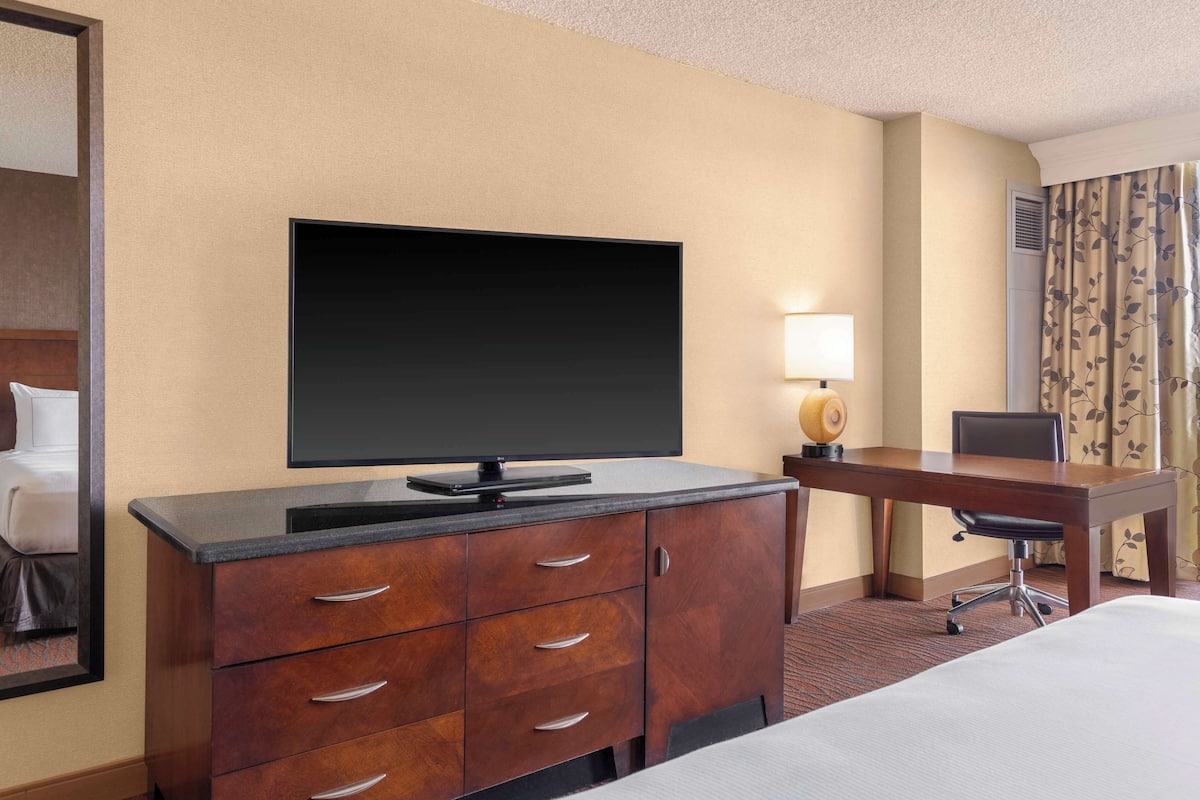 Quarto de hotel com cama, escrivaninha, cadeira, televisão de tela plana, cômoda e abajur.