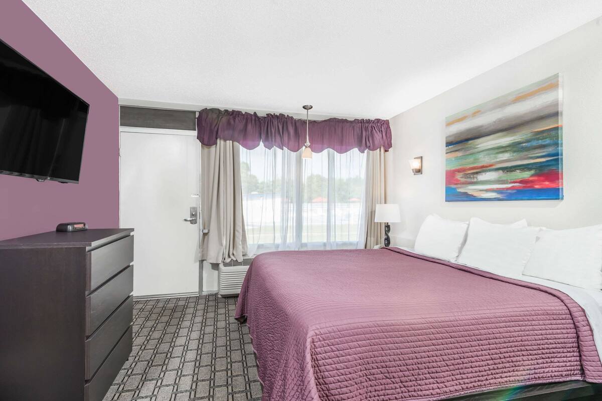 A hotel room with a bed, a dresser, a flat-screen TV, a painting on the wall, and a window with curtains.
