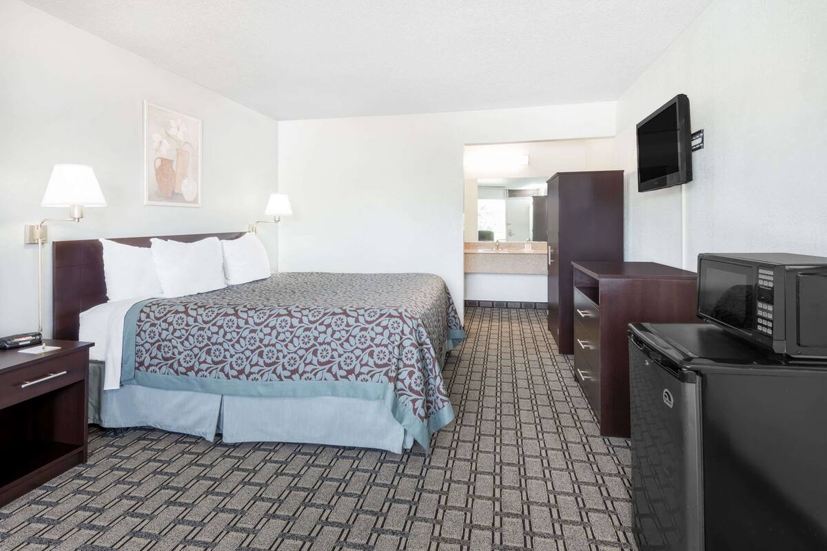 A hotel room with a bed, bedside lamps, a television, a refrigerator, and a bathroom visible through an open door.