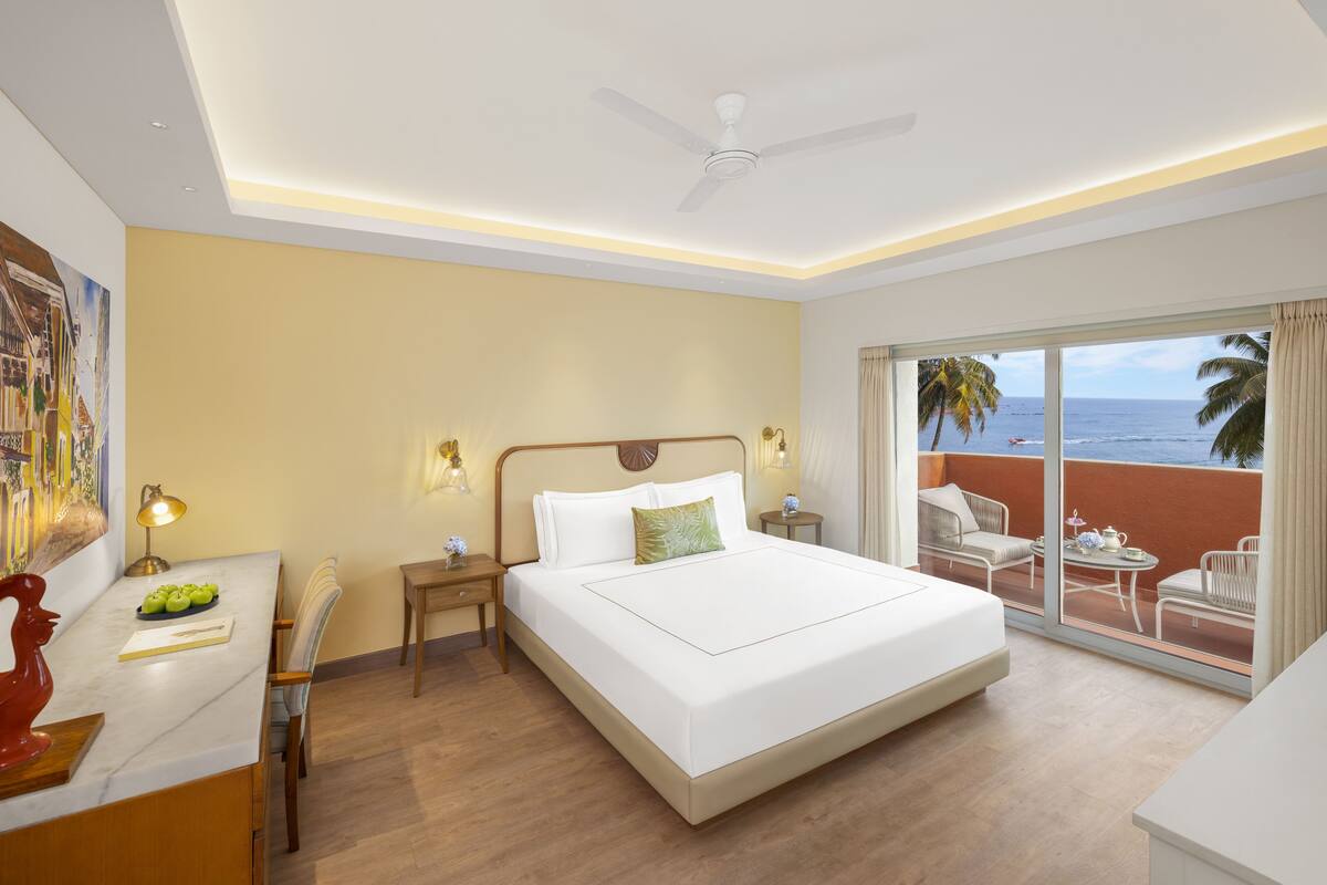 Une chambre d’hôtel avec un grand lit, un bureau avec une lampe, une chaise, une table de chevet et une vue sur l’océan par une porte coulissante.