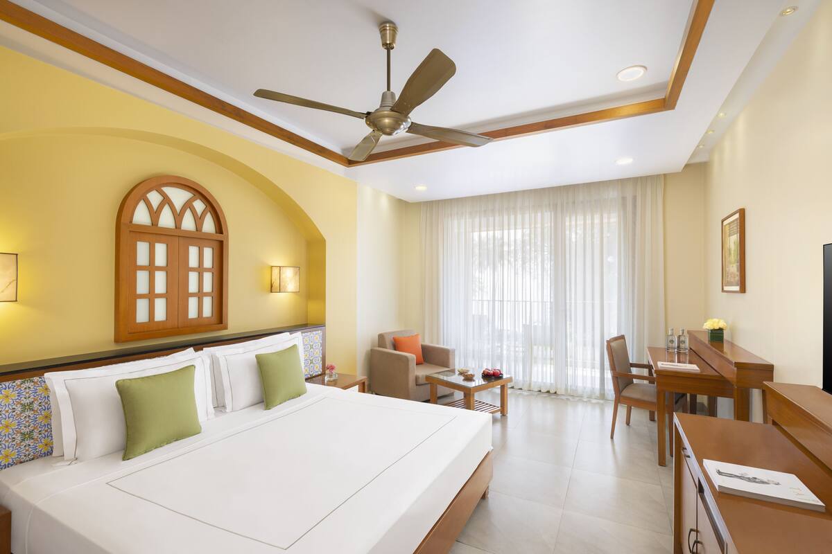Une chambre d’hôtel avec un grand lit, un ventilateur de plafond, une table à manger avec des chaises et une fenêtre avec des rideaux.