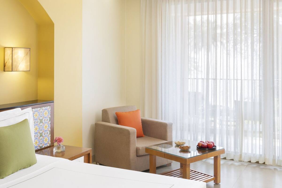 Une chambre d’hôtel avec un lit, une chaise, une petite table avec des fruits et une grande fenêtre avec des rideaux légers.