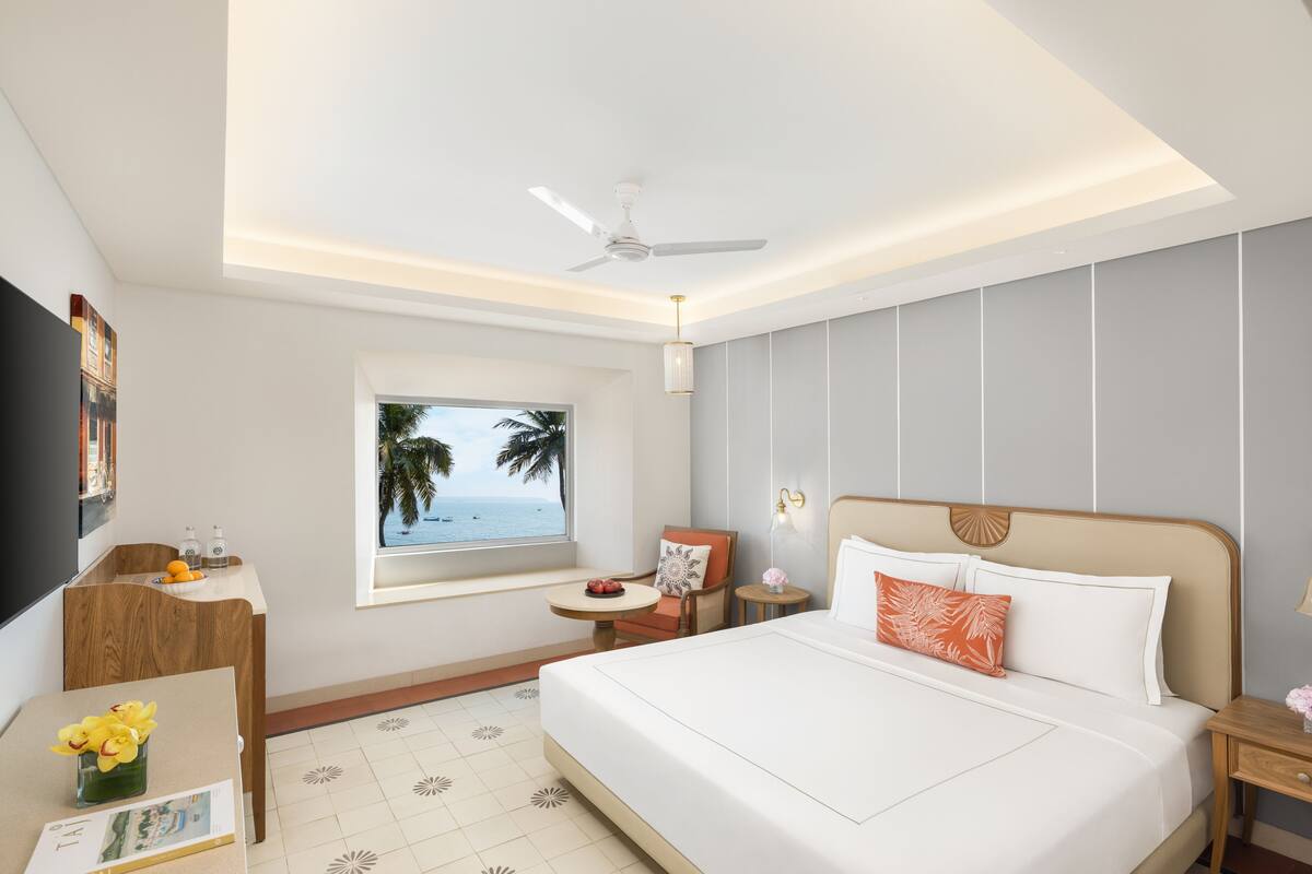 Une chambre d’hôtel avec un grand lit, un bureau avec une chaise, une petite table, un ventilateur de plafond, une fenêtre donnant sur des palmiers et un téléviseur fixé au mur.