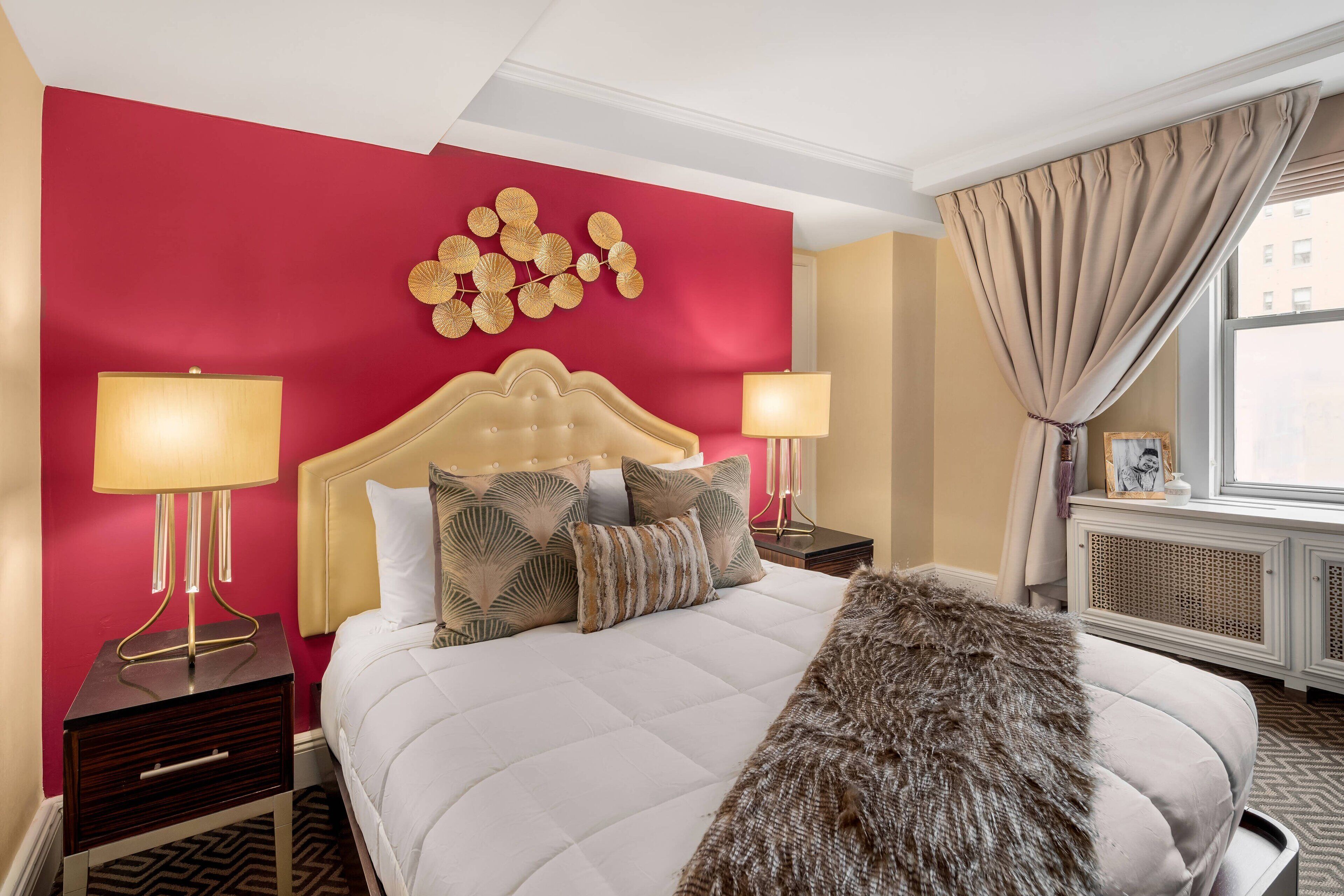 A hotel room with a red accent wall, a tufted headboard, a large bed with patterned pillows, two bedside lamps, a fur throw, and a view of the city through a window with beige curtains.