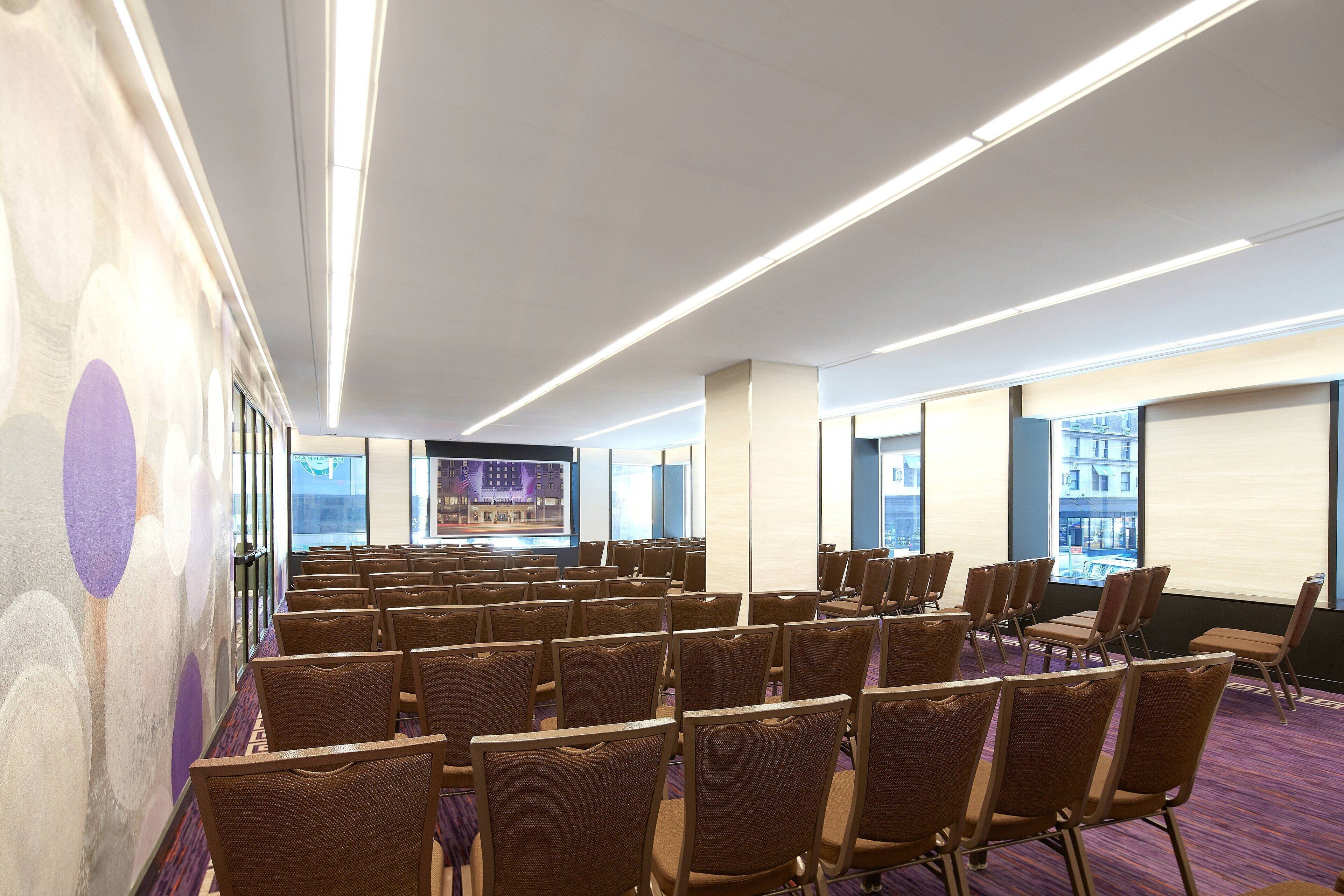A conference room with rows of chairs, a large screen, and a city view through the windows.