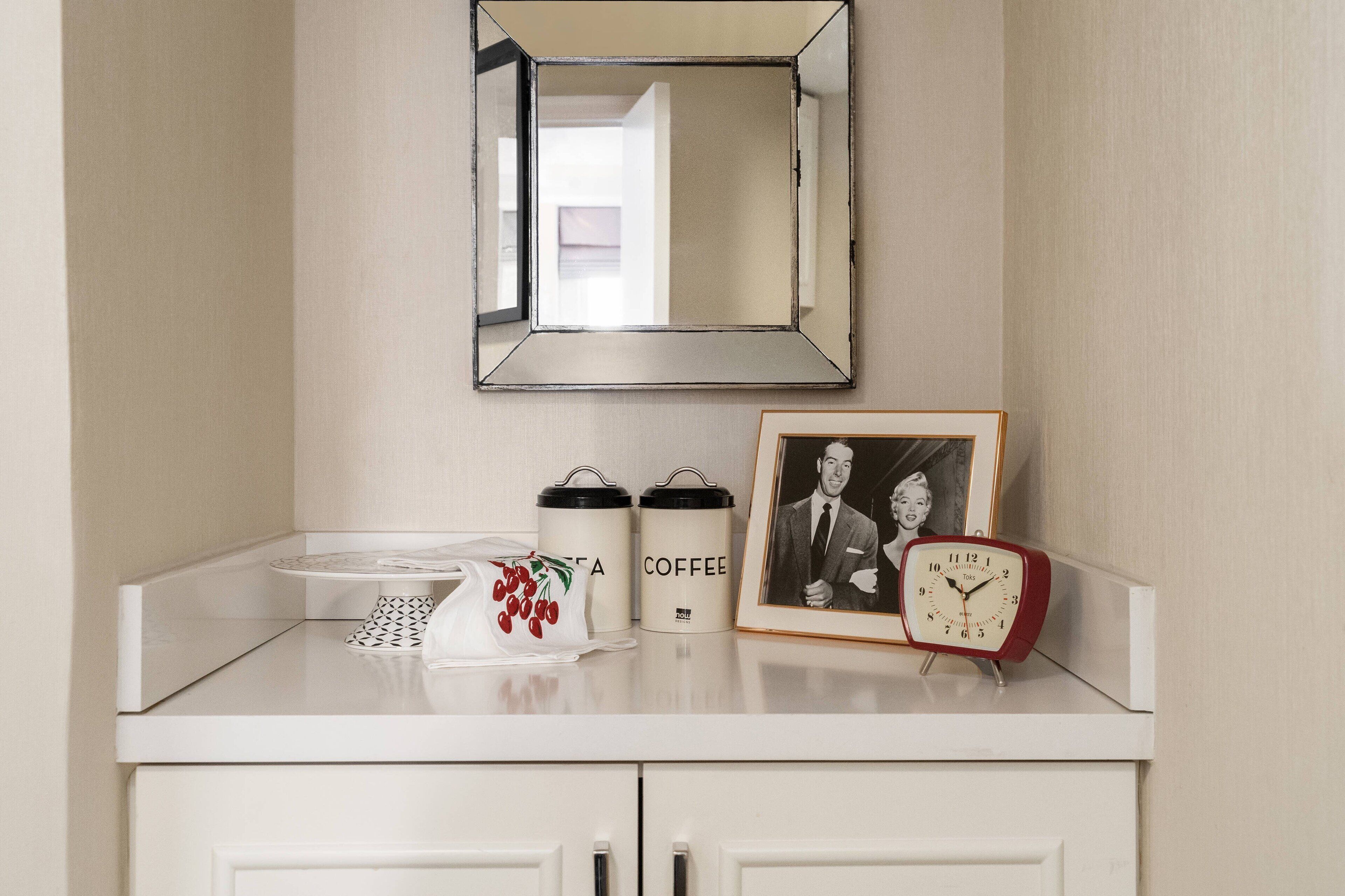 Et badeværelsesbord med spejl, to kaffekander, et indrammet foto og en rød alarmklokke.