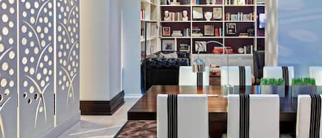 Un comedor moderno con una mesa de madera y sillas blancas tapizadas, una estantería llena de libros y objetos decorativos, y una alfombra estampada.