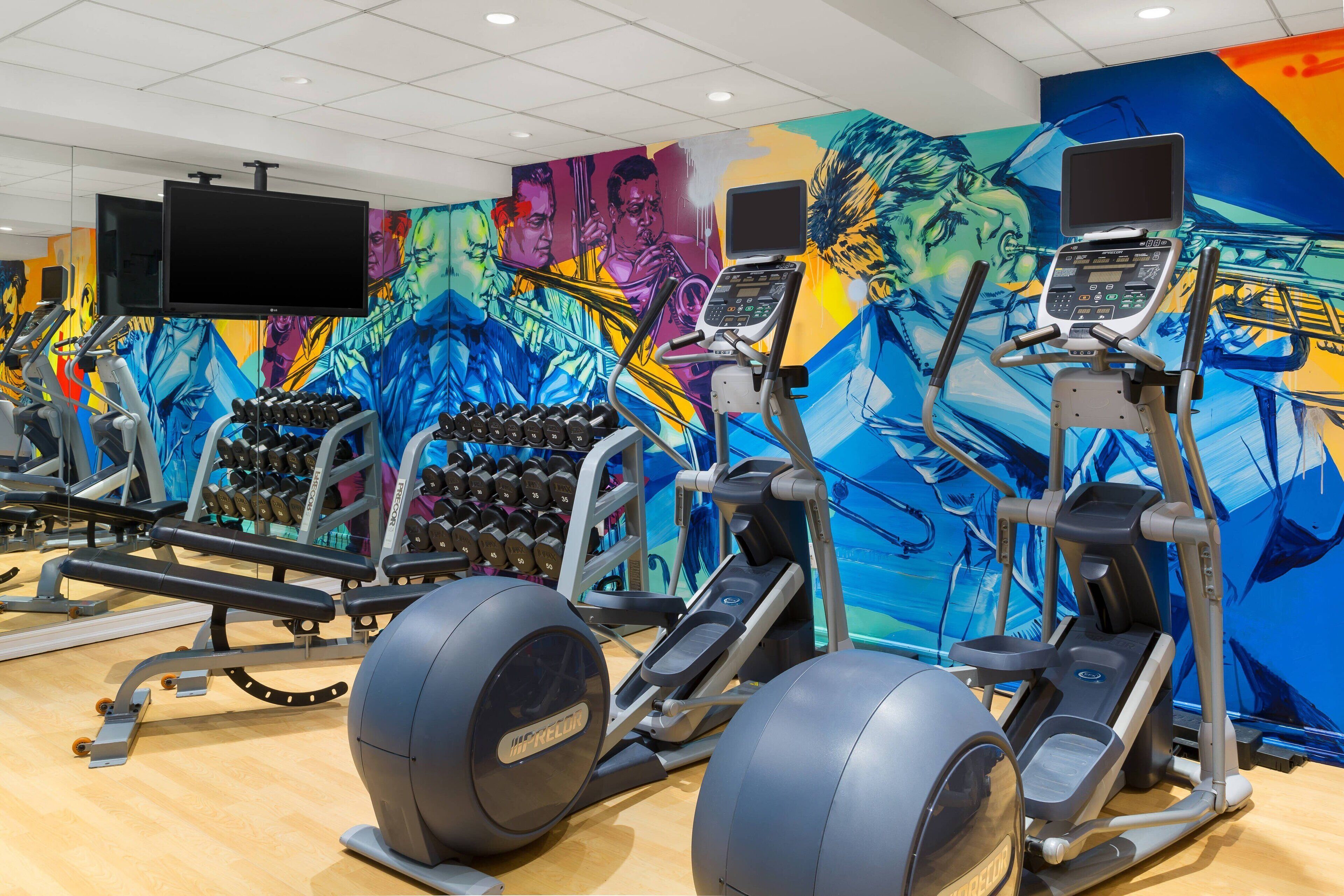 A gym with exercise equipment, a wall with a colorful mural, and a television mounted on the wall.