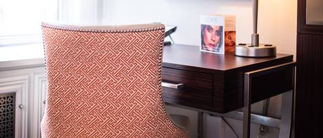 A hotel room with a patterned armchair, a desk with a lamp, and a framed picture on the wall.