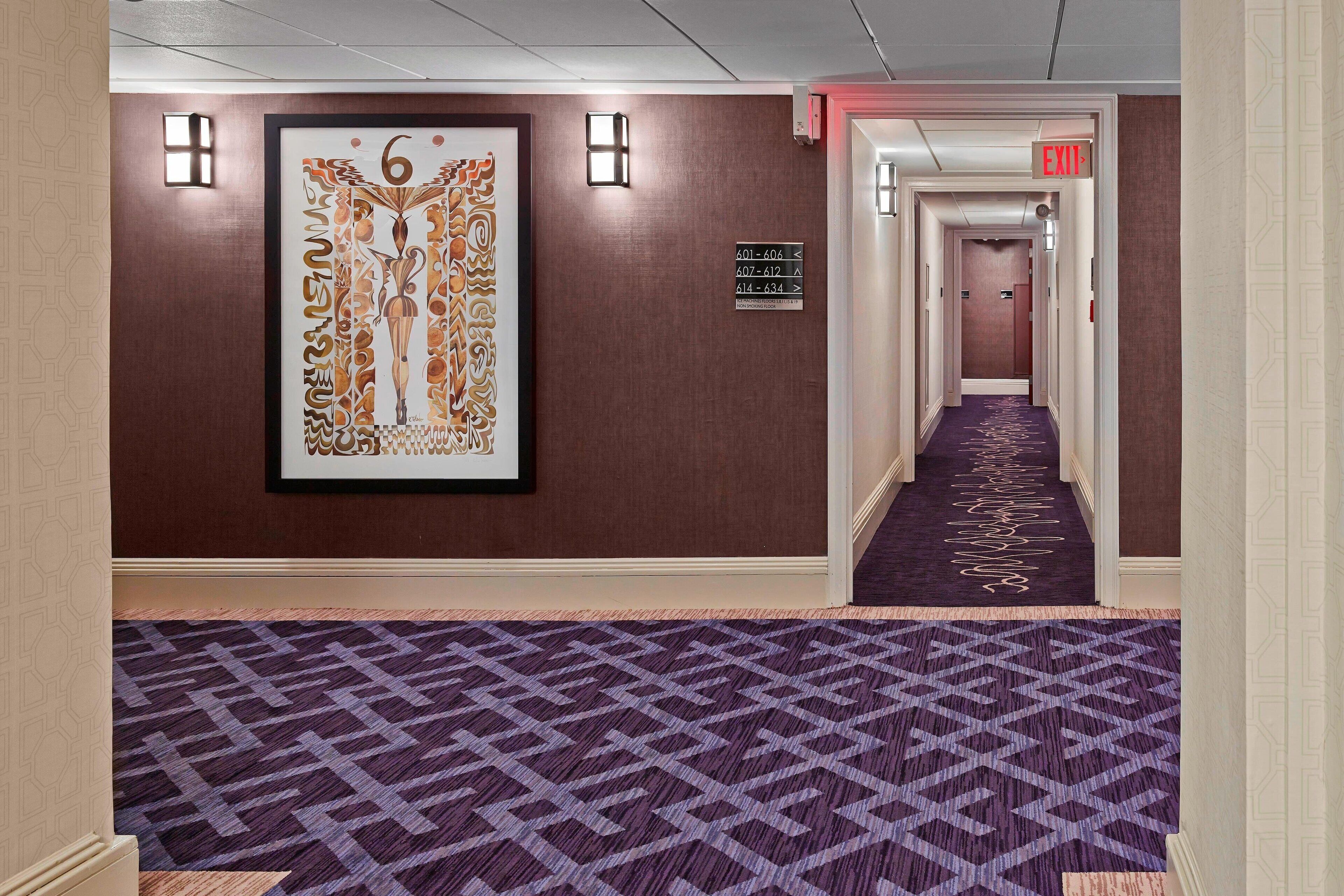 A hotel hallway with a patterned carpet, a framed artwork on the wall, and directional signs.