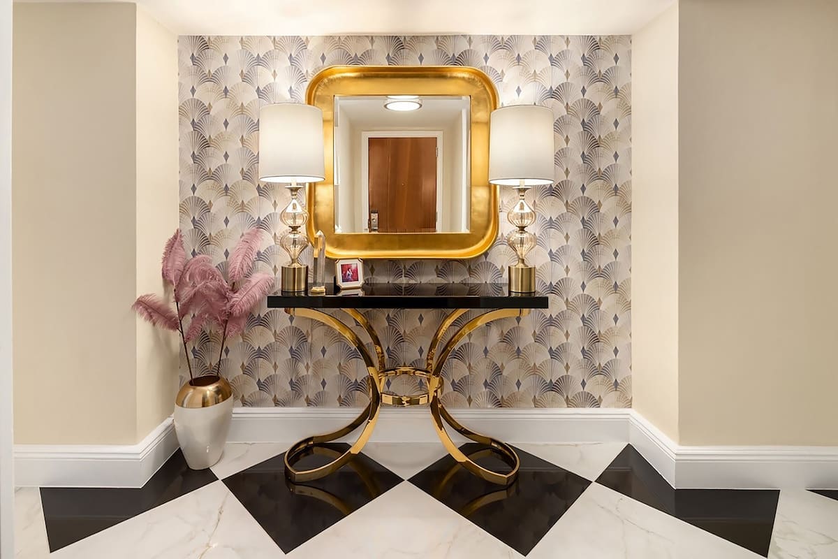 Un baño con tocador, espejo, dos lámparas de pared, un jarrón decorativo y papel tapiz estampado.