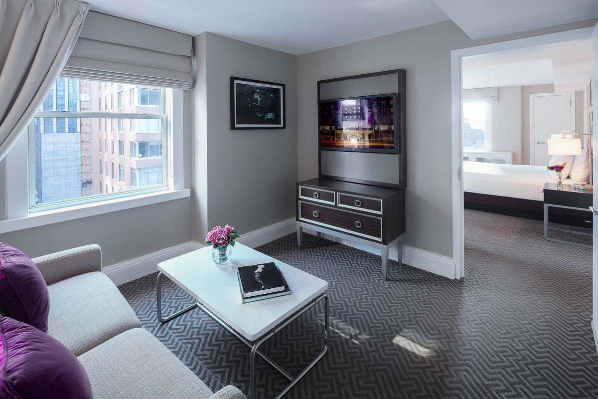 A hotel room with a bed, a TV, a desk with a book and flowers, and a view of buildings through the window.