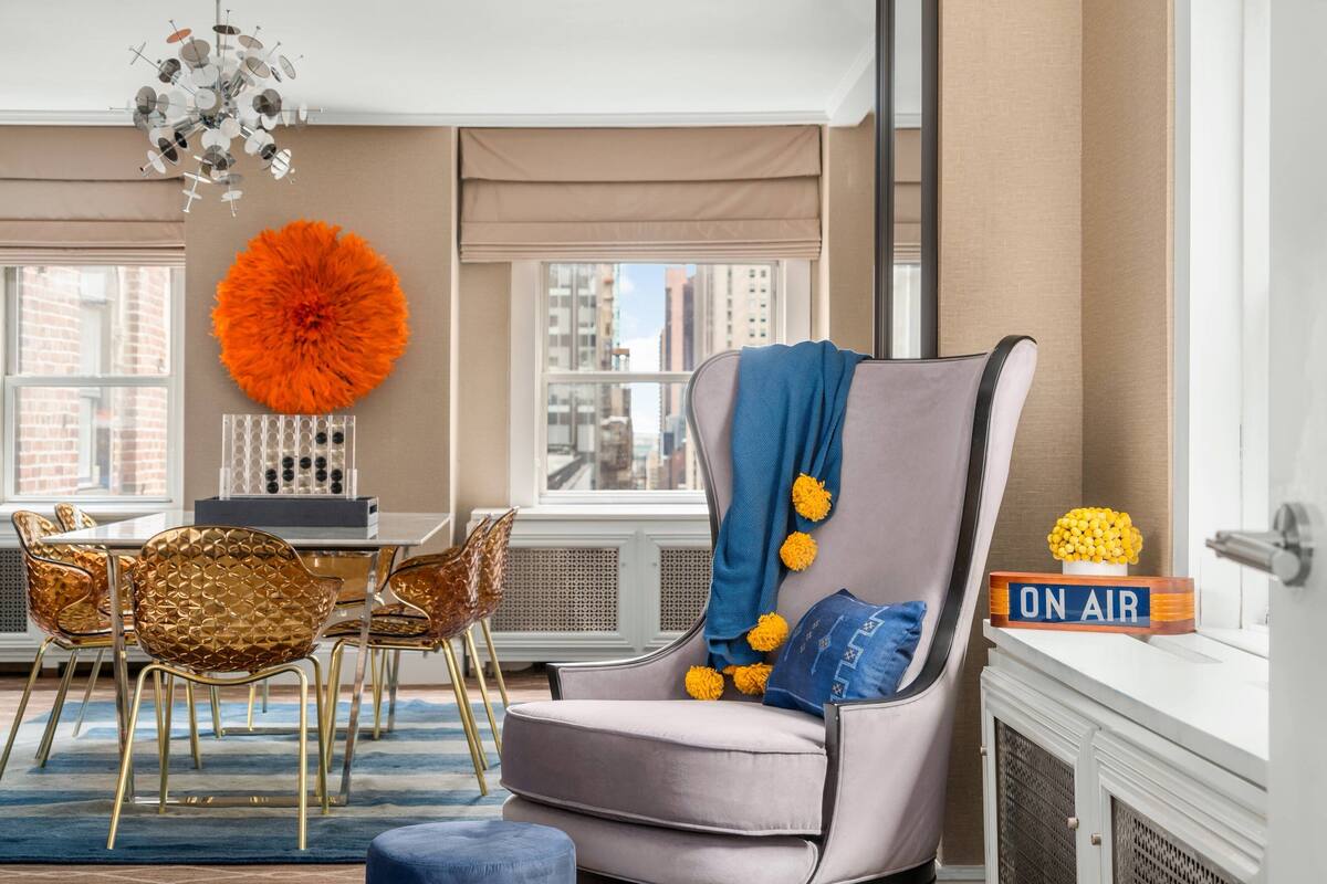 A modern living room with a grey armchair, a dining table with golden chairs, a large orange decorative piece, and a view of the city through large windows.